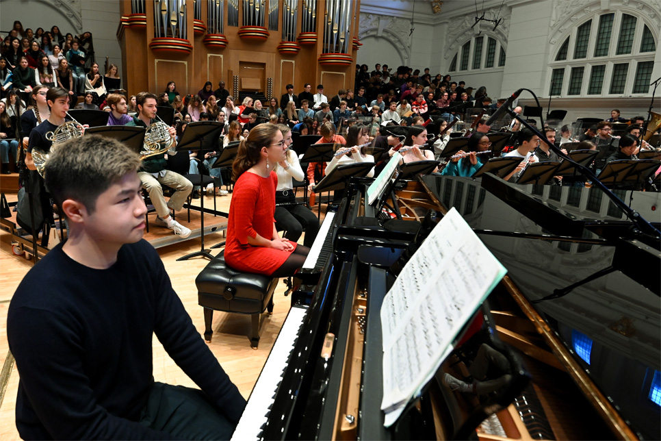 Two pianist students, sitting at two pianos, with a full orchestra and choir behind them, on stage in a performance space.